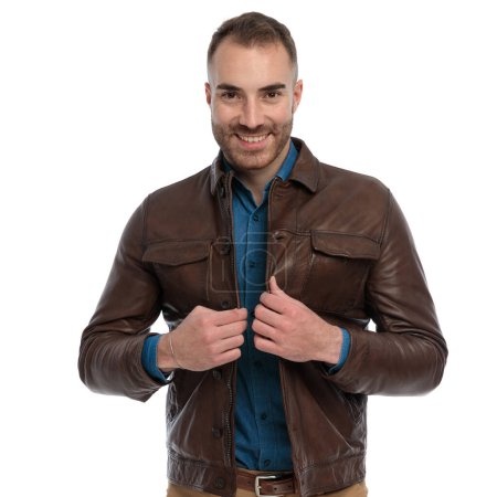 Foto de Portrait of happy young man smiling and closing brown leather jacket in front of white background in studio - Imagen libre de derechos
