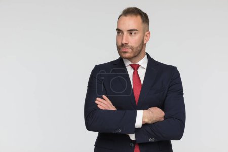 Foto de Confident businessman in elegant suit looking to side while crossing arms and posing in front of grey background in studio - Imagen libre de derechos