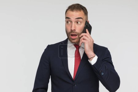 Foto de Surprised young man having a phone conversation and looking to side, posing in a shocked manner on grey background - Imagen libre de derechos