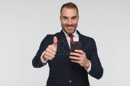 Téléchargez les photos : Confident businessman reading emails and making thumbs up gesture, smiling and posing on grey background in studio - en image libre de droit