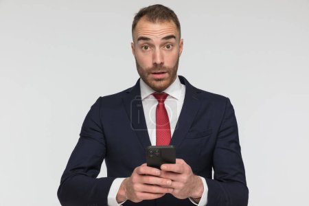 Téléchargez les photos : Shocked man in suit holding phone in his hands and reading emails, being surprised and posing in front of grey background in studio - en image libre de droit