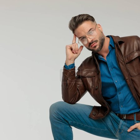 Foto de Portrait of sexy casual man putting hand in pocket and touching head, leaning on one leg, wearing eyeglasses against gray studio background - Imagen libre de derechos