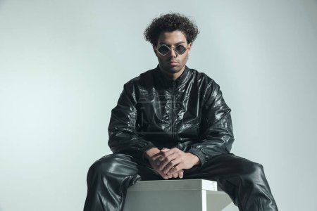 Téléchargez les photos : Sexy fashion man with curly hair posing in a cool way while wearing leather outfit and sunglasses while sitting on grey background in studio - en image libre de droit