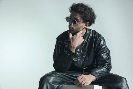 Photo for Confident cool fashion man with sunglasses looking to side and touching chin, thinking and sitting on grey background in studio - Royalty Free Image