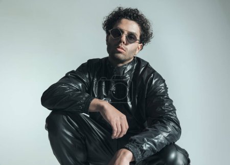 Photo for Young fashion man with sunglasses wearing leather jacket and crouching while posing in front of grey background in studio - Royalty Free Image