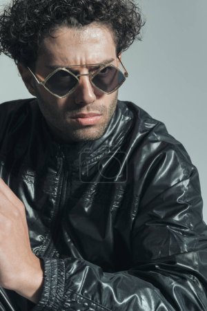 Téléchargez les photos : Arabic guy with curly hair wearing sunglasses and leather jacket posing while looking away on grey background - en image libre de droit