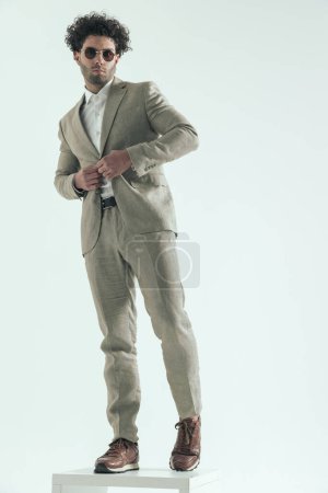 Foto de Full body picture of sexy turkish businessman with sunglasses buttoning suit and posing in front of grey background in studio - Imagen libre de derechos