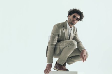 Photo for Side view of handsome curly hair lebanese guy with sunglasses crouching and holding elbow above knee on grey background - Royalty Free Image