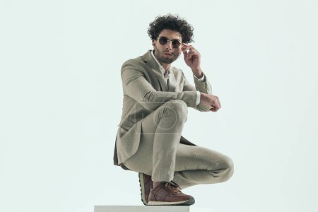 Photo for Fashion man in suit with curly hair adjusting sunglasses and posing with elbow above head in front of grey background in studio - Royalty Free Image