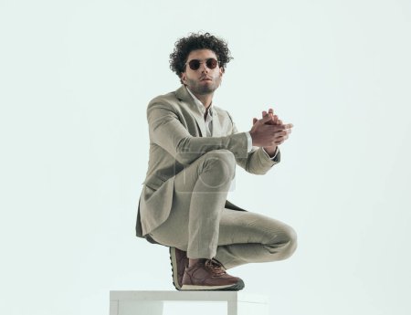 Foto de Full body picture of lebanese fashion model with round glasses and curly hair crouching with elbow on knee and rubbing palms on grey background - Imagen libre de derechos