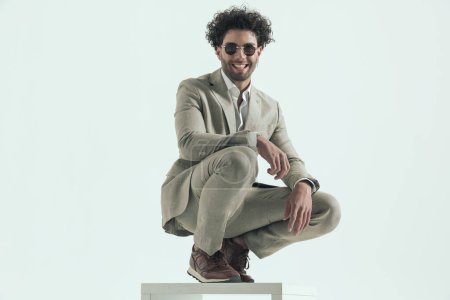 Photo for Curly hair man in suit with sunglasses laughing and crouching with elbow on knee in front of grey background in studio - Royalty Free Image