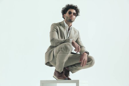 Photo for Curly hair guy in elegant suit holding elbow on knee and crouching while posing in front of grey background in studio - Royalty Free Image
