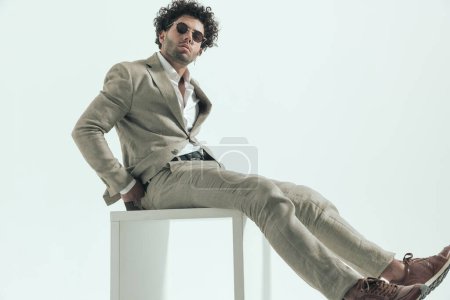 Photo for Sexy businessman with curly hair and glasses adjusting suit while sitting in front of grey background in studio - Royalty Free Image