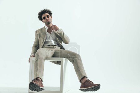 Téléchargez les photos : Curly hair man with sunglasses adjusting shirt collar and posing while sitting in front of grey background in studio - en image libre de droit