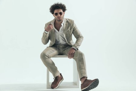 Téléchargez les photos : Curly hair arabic guy with sunglasses adjusting shirt collar and sitting in front of grey background in studio - en image libre de droit