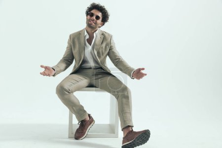 Foto de Happy sexy arabic man with sunglasses smiling and opening arms while sitting in front of grey background in studio - Imagen libre de derechos