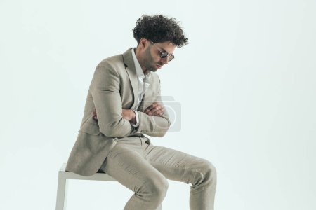Foto de Side view of curly hair man with crossed arms sitting and looking down in front of grey background in studio - Imagen libre de derechos