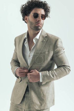 Téléchargez les photos : Handsome lebanese man with curly hair fixing and opening suit, wearing sunglasses and looking to side while posing on grey background in studio - en image libre de droit