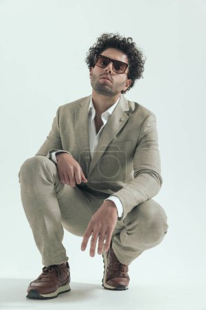 Photo for Confident greek man with glasses posing with elbows on knees in front of grey background while crouching in studio - Royalty Free Image