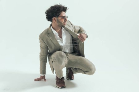 Photo for Picture of attractive turkish man looking to side while pulling suit and touching floor, crouching in front of grey background in studio - Royalty Free Image
