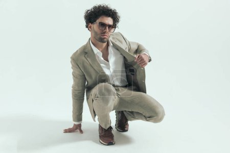 Téléchargez les photos : Handsome greek guy with sunglasses adjusting and pulling suit while crouching in front of grey background in studio - en image libre de droit