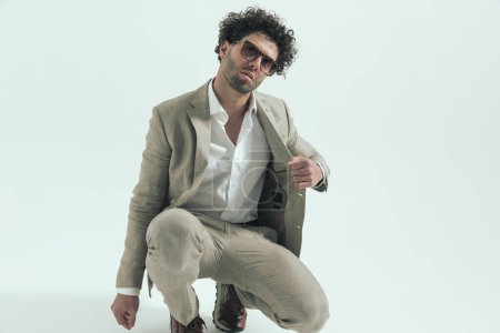 Photo for Cool fashion arabic man with sunglasses crouching and pulling suit in front of grey background in studio - Royalty Free Image