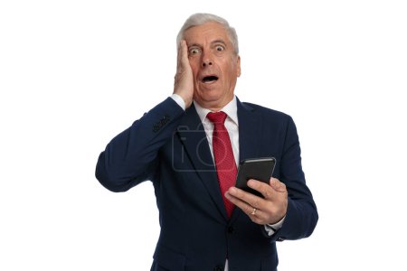 Foto de Old businessman slapping his face feeling utterly shocked while holding his mobile phone - Imagen libre de derechos