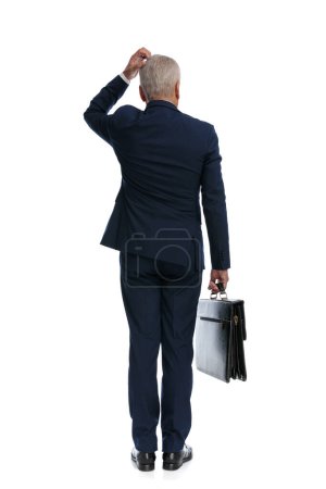 Photo for Full body picture and rear view of an old business man scratching his head, pondering and important decision and holding a briefcase - Royalty Free Image