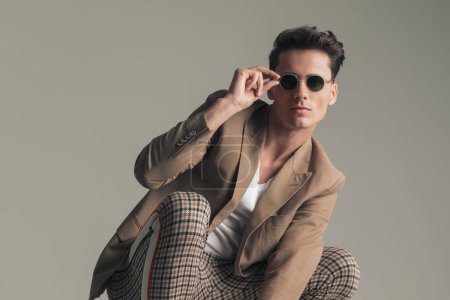 Foto de Attractive man with dark hair crouching and posing while adjusting sunglasses in front of grey backgroudn in studio - Imagen libre de derechos