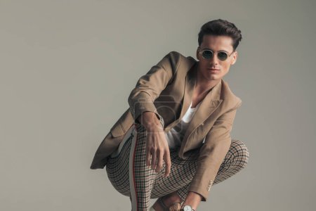 Foto de Happy man with sunglasses wearing beige jacket and plaid pants and crouching in front of grey background in studio - Imagen libre de derechos