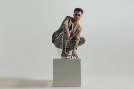 Téléchargez les photos : Casual man with dark hair wearing beige jacket and sunglasses and crouching on top of white box in front of grey background - en image libre de droit