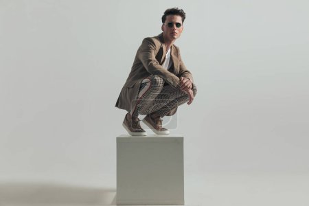 Photo for Cool young guy with sunglasses in casual outfit standing on top of white box and posing with elbows on knees in front of grey background - Royalty Free Image