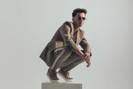 Foto de Cool fashion model with plaid pants and beige jacket crouching on top of white box and posing with elbows above knees in front of grey background in studio - Imagen libre de derechos