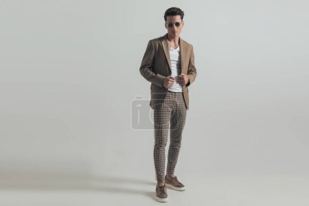 Foto de Sexy man with dark hair wearing plaid pants and adjusting jacket while posing in front of grey background in studio, full length - Imagen libre de derechos