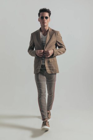 Téléchargez les photos : Full length picture of young man with dark hair buttoning beige jacket and walking with confidence in front of grey background in studio - en image libre de droit