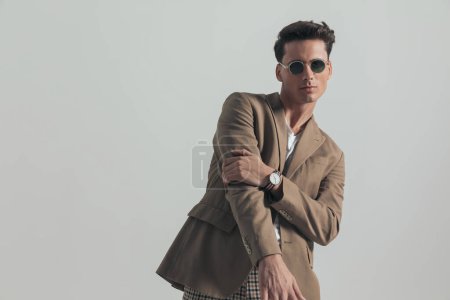 Téléchargez les photos : Portrait of cool casual man with beige jacket and sunglasses holding arms in fashion pose and posing in front of grey background in studio - en image libre de droit