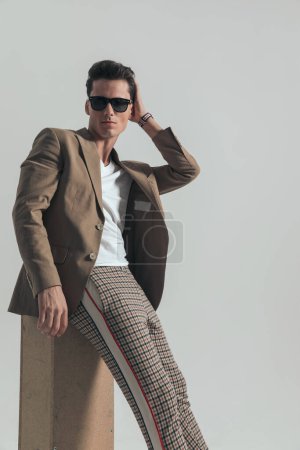Foto de Stylish young guy in casual outfit adjusting hairstyle while laying on a box in front of grey background in studio - Imagen libre de derechos