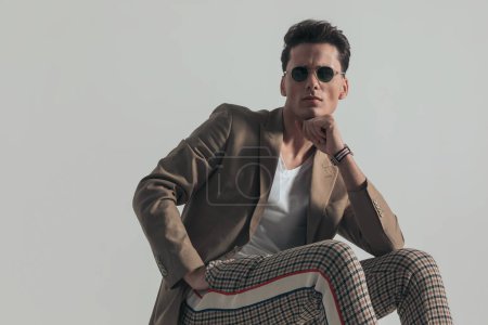 Photo for Cool man with beige jacket and plaid pants holding hands in pocket, holding elbow on knee and touching chin, thinking on grey background in studio - Royalty Free Image