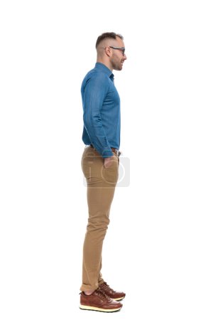 Foto de Handsome man in denim shirt holding hands in pockets and waiting in line in front of white background in studio - Imagen libre de derechos
