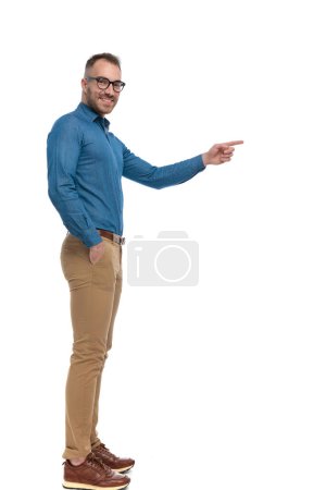 Photo for Side view of handsome guy standing in line with hand in pocket and pointing finger while smiling on white background in studio - Royalty Free Image