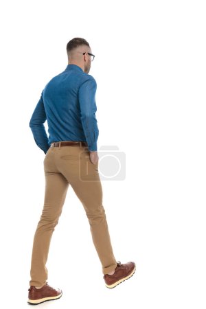 Foto de Rear view of handsome casual guy in denim shirt walking with  hands in pockets on white background in studio - Imagen libre de derechos