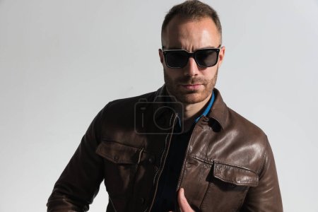 Foto de Portrait of casual man with sunglasses adjusting brown leather jacket and posing in a cool way on grey background in studio - Imagen libre de derechos