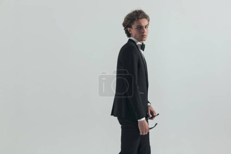 Téléchargez les photos : Side view of caucasian young man with blonde curly hair holding glasses in front of grey background - en image libre de droit