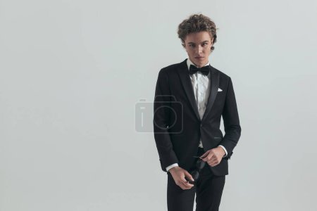 Foto de Portrait of handsome curly hair man holding glasses and posing in front of grey background while wearing elegant black tuxedo - Imagen libre de derechos