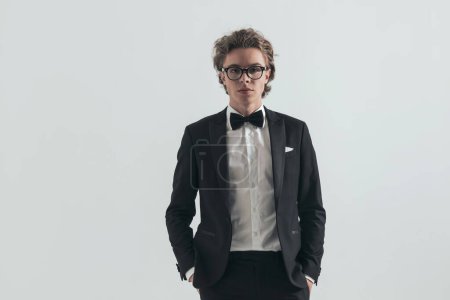 Téléchargez les photos : Portrait of elegant man in tuxedo with eyeglasses holding hands in pockets and posing in front of grey background in studio - en image libre de droit