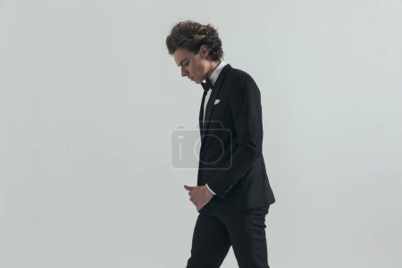 Téléchargez les photos : Side view of blonde man with curly hair in black tuxedo walking and looking down in front of grey background in studio - en image libre de droit