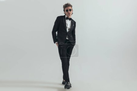 Téléchargez les photos : Curly hair man wearing black elegant tuxedo and posing with hands in pockets in front of grey background - en image libre de droit