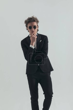 Photo for Cool elegant best man in tuxedo with curly hair touching mouth and posing with arms crossed in front of grey background in studio - Royalty Free Image