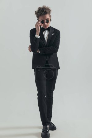 Téléchargez les photos : Confidetn man with curly hair wearing black tuxedo and adjusting sunglasses while posing in front of grey background in studio - en image libre de droit