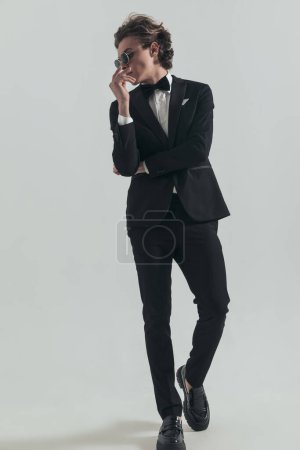 Photo for Fashion young man in elegant tuxedo looking to side and posing with arms to mouth in a cool way on grey background - Royalty Free Image